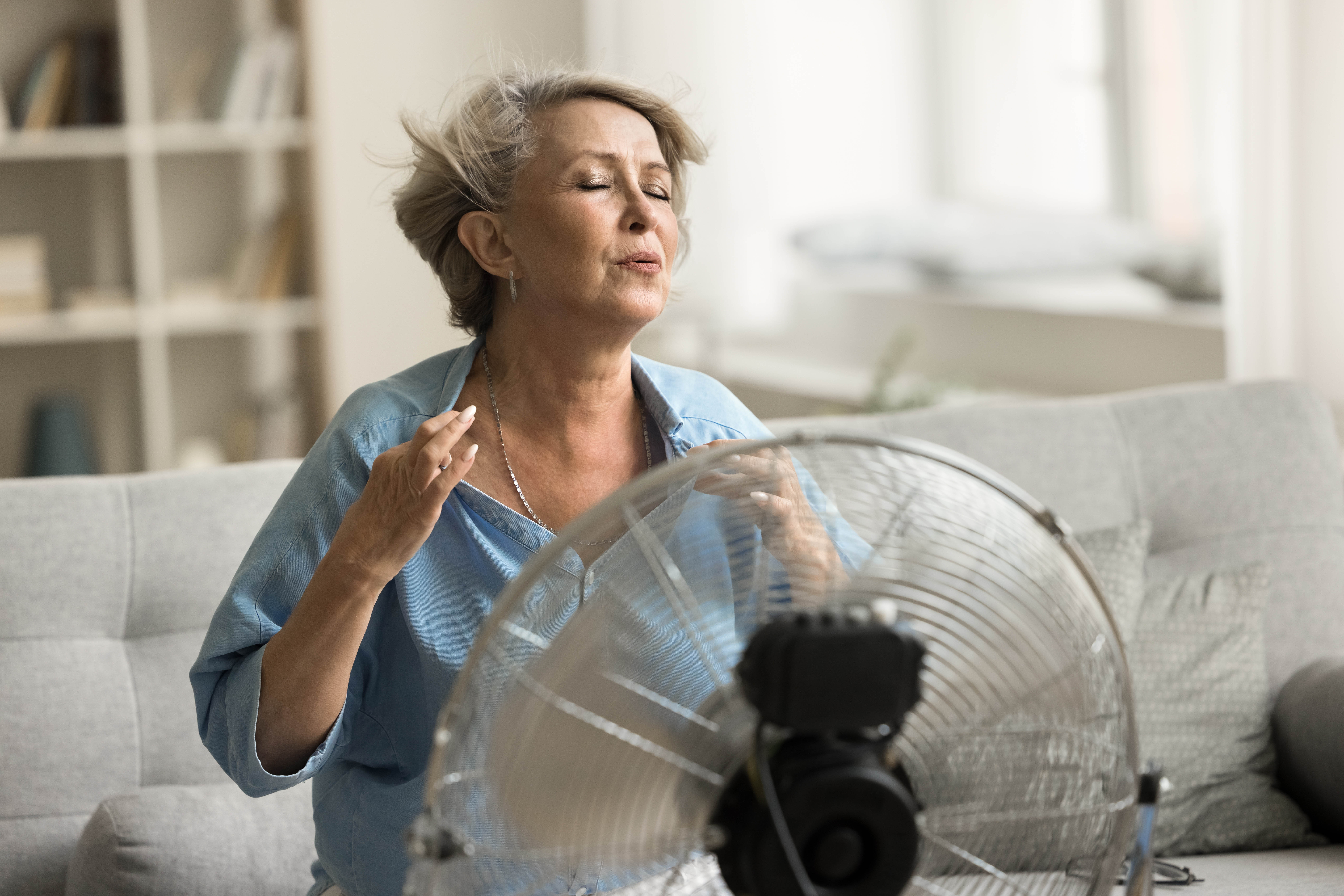 Older woman sitting on her couch, with a fan right in front of her trying to cool down Older woman sitting on her couch, with a fan right in front of her trying to cool down